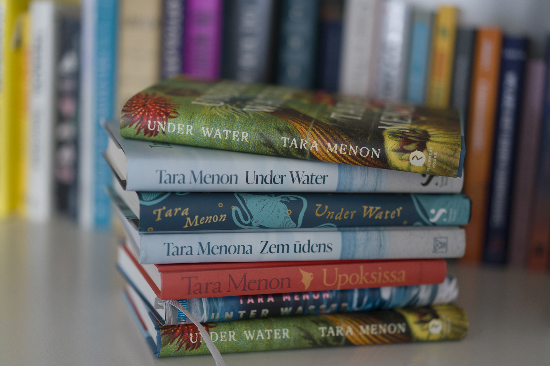 A stack of various editions of Tara Menon's novel "Under Water," published in several languages