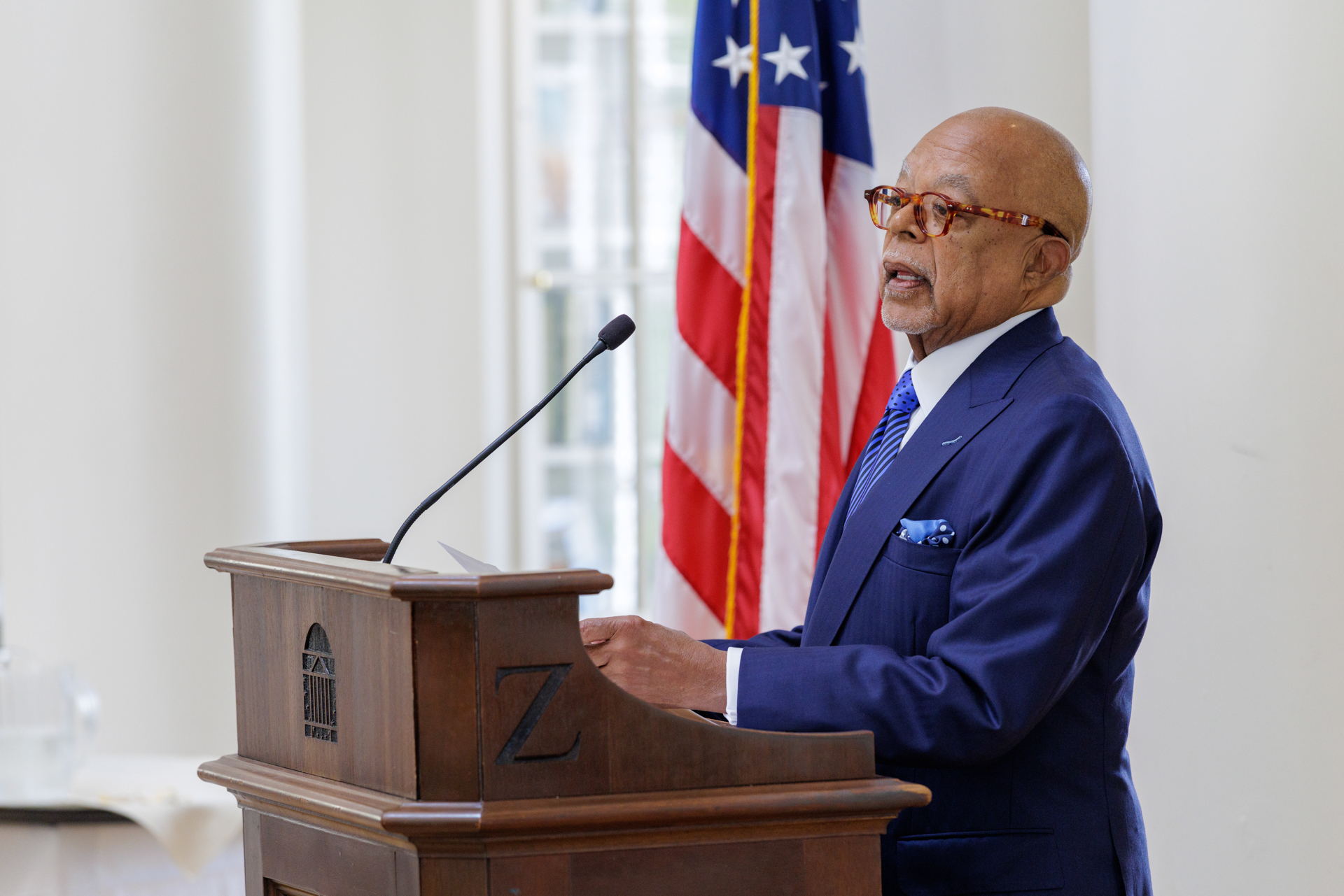 Henry Louis Gates Jr. speaks at a podium with an American flag in the background.