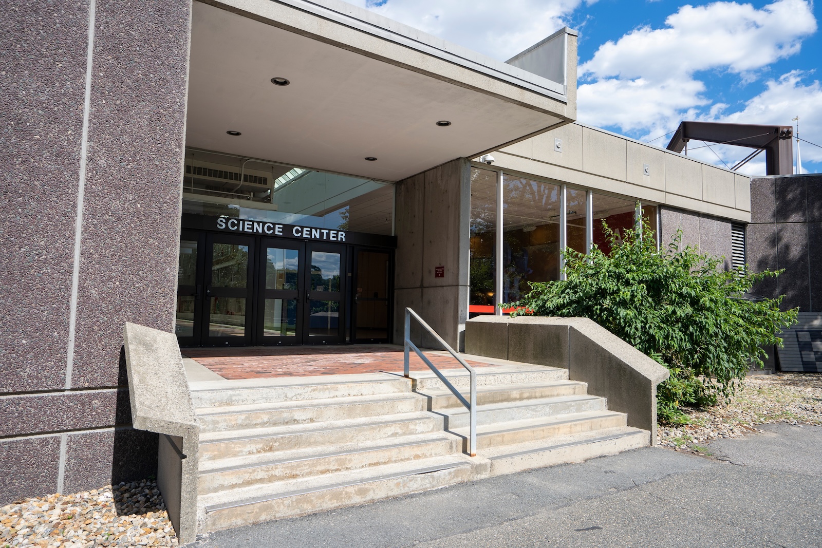 The concrete steps and glass entrance of the Harvard Science Center under a bright, partly cloudy sky.