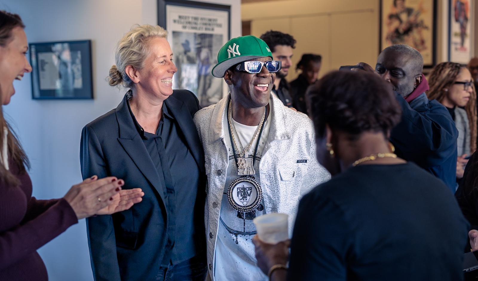 Flavor Flav, wearing a white bedazzled jacket, green New York hat, and his signature clock pendant, shares a laugh with a smiling guest. The scene captures the "celebratory energy" of the listening party hosted at the Hutchins Center for African & African American Research.