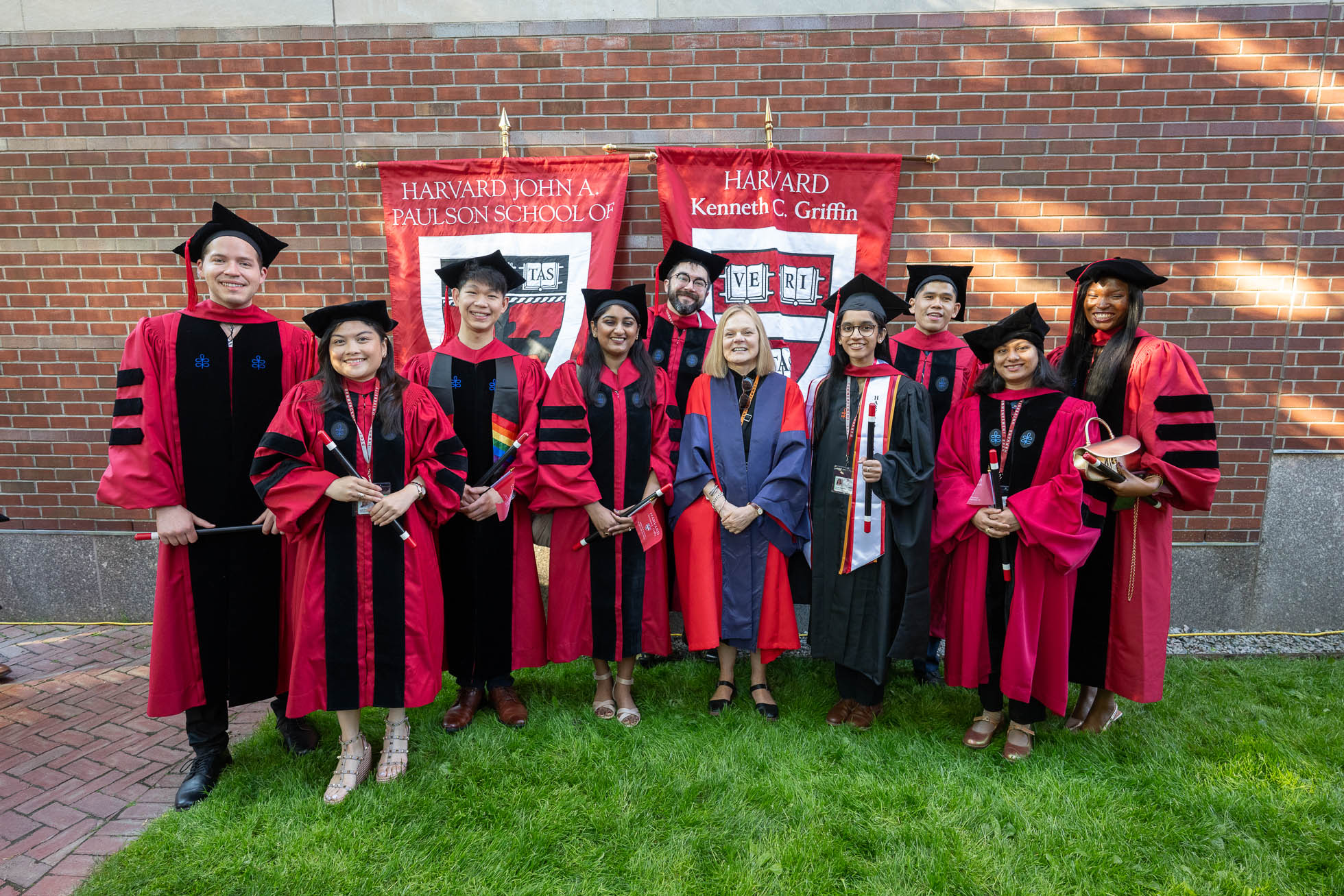 Emma Dench, Dean of the Harvard Kenneth C. Griffin Graduate School of Arts and Sciences, in red and blue at center, posed with graduates outside Sanders Theatre.