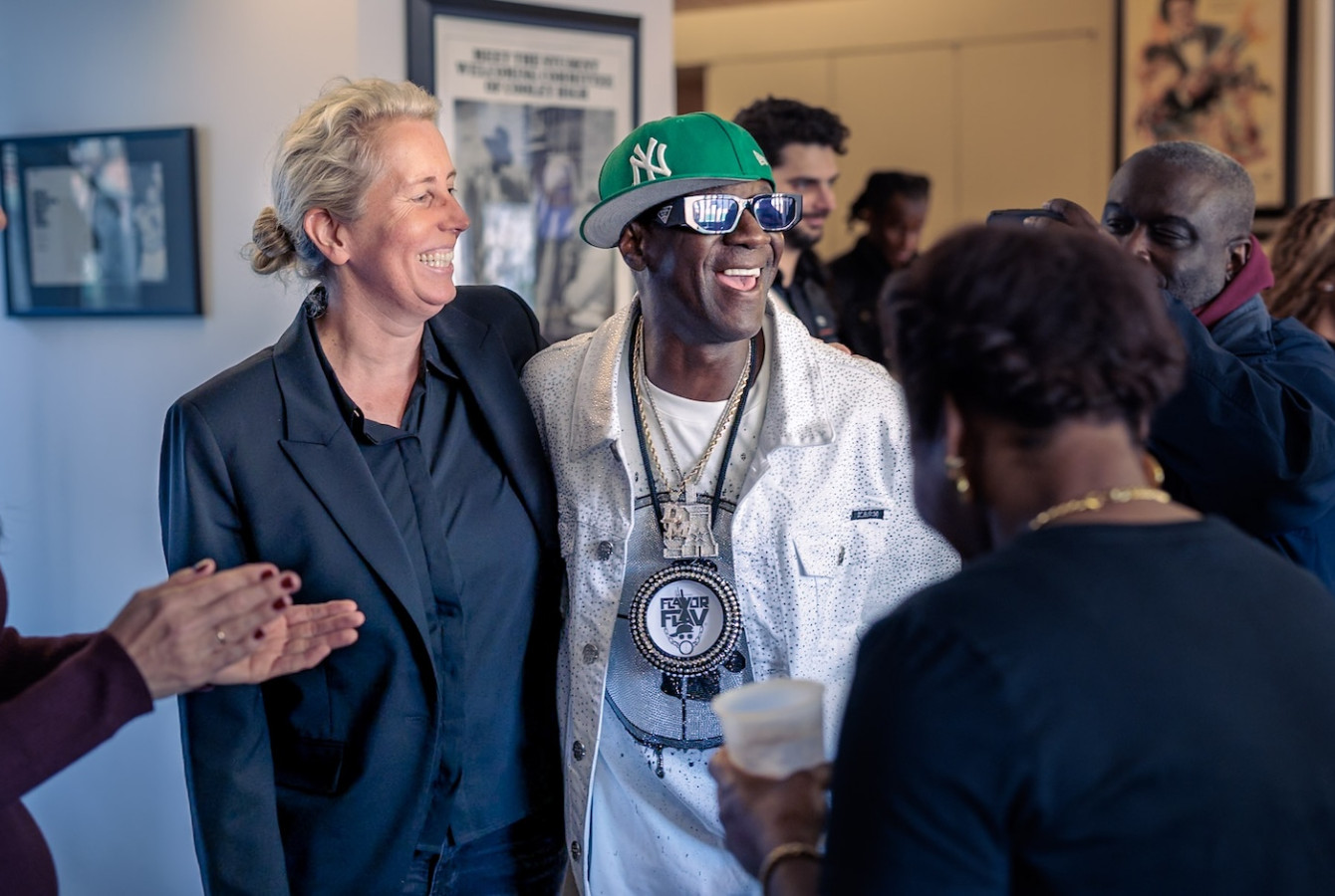 Flavor Flav, wearing a white bedazzled jacket, green New York hat, and his signature clock pendant, shares a laugh with a smiling guest. The scene captures the "celebratory energy" of the listening party hosted at the Hutchins Center for African & African American Research.
