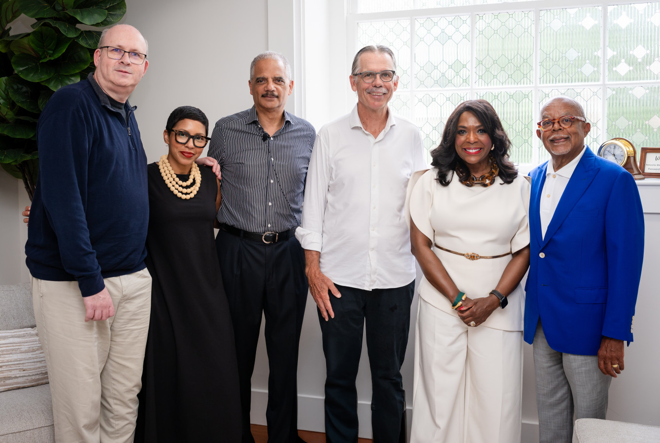 Group photo of six speakers and hosts standing side by side indoors, dressed in smart casual attire, smiling at the camera in front of a window.