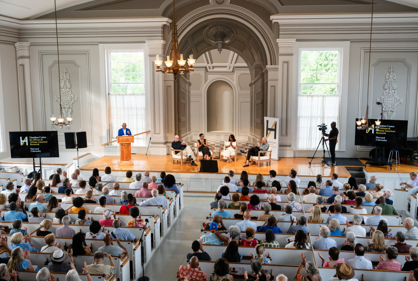 High-angle wide view of a crowded hall with an audience seated in pews facing a wooden stage. On stage, a host stands at a podium on the left while four panelists sit in armchairs across the center, with a videographer standing on the right.