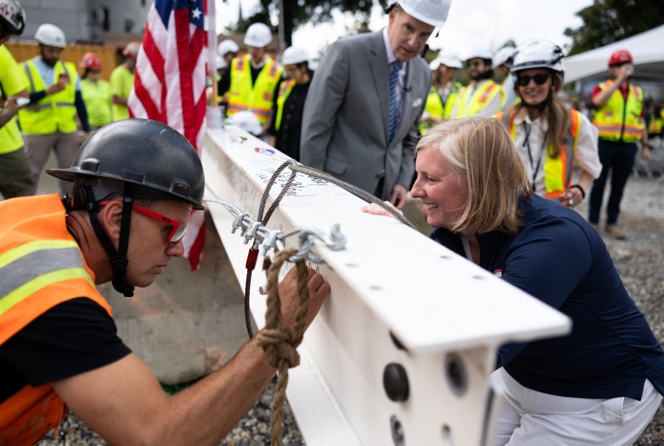 A man in a hard hat and orange vest signs a beam while Dean Hopi Hoekstra looks on