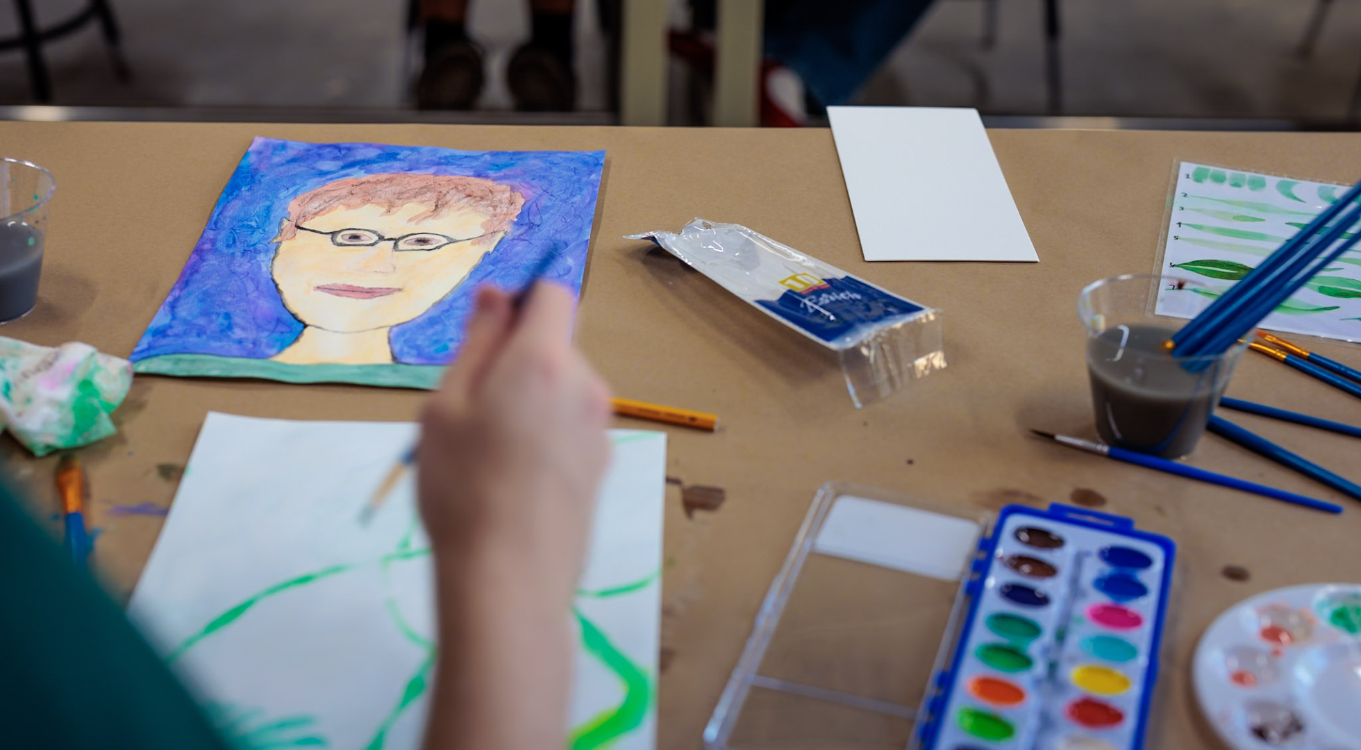 A student’s hand working on a watercolor painting, with a completed work and paint supplies on the table
