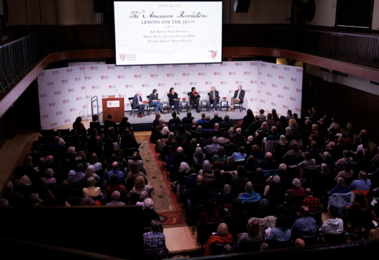 Panelists on a stage in front of a Harvard Kennedy School step and repeat logo in front of an audience