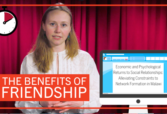 Student Gabriella Fleischman in front of a red curtain with graphics reading “The Benefits of Friendship” and ”Economic and and Psychological Returns to Social Relationships: Alleviating Constraints to Network Formation in Malawi”