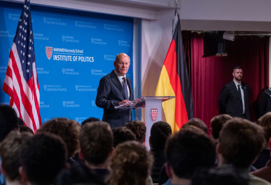 Olaf Scholz speaks at the John F. Kennedy Jr. Forum, on stage at a podium, flanked by American and German flags