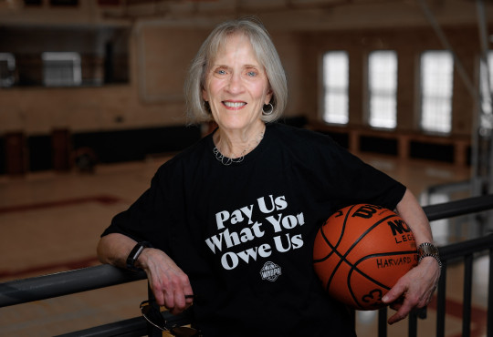 Claudia Goldin holding a basketball and wearing a black T-shirt that reads ”Pay Us What You Owe Us”