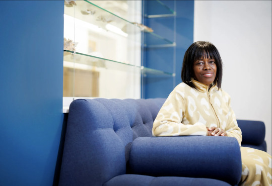 Mandë Holford is seated on a modern blue sofa, smiling warmly at the camera. Behind her, a glass display case showcases various marine mollusk shells