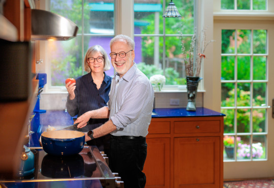 Professor Dan Lieberman and Tonia Prescott in their kitchen. Lieberman stirs a pot on the stove and Prescott holds a tomato.