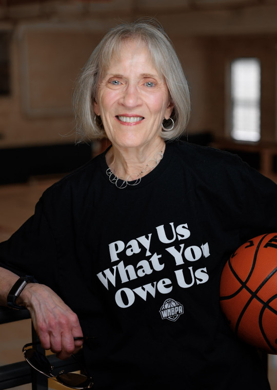 Claudia Goldin holding a basketball and wearing a black T-shirt that reads ”Pay Us What You Owe Us”