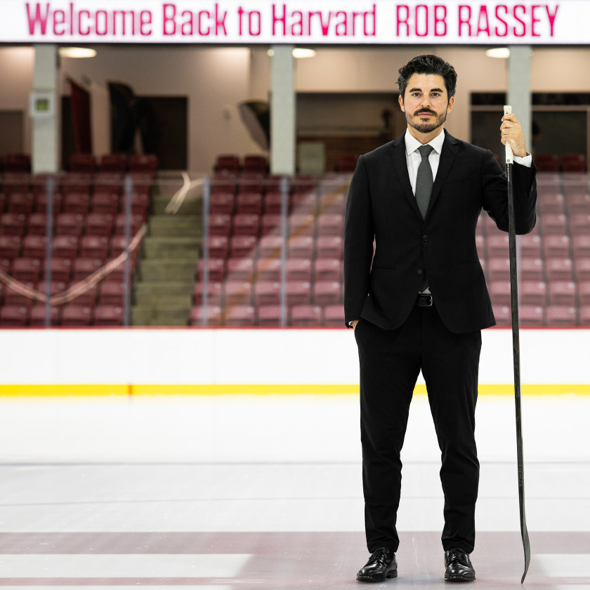 Men’s ice hockey coach Ron Rassey wearing a suit and holding a hockey stick at center ice in the Bright-Landry Hockey Center