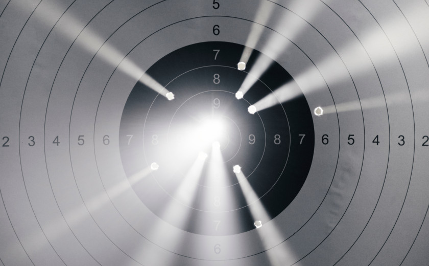 A black and white shooting target with several bullet holes. Beams of bright, ethereal light shine through each puncture, creating a glowing effect against the paper rings
