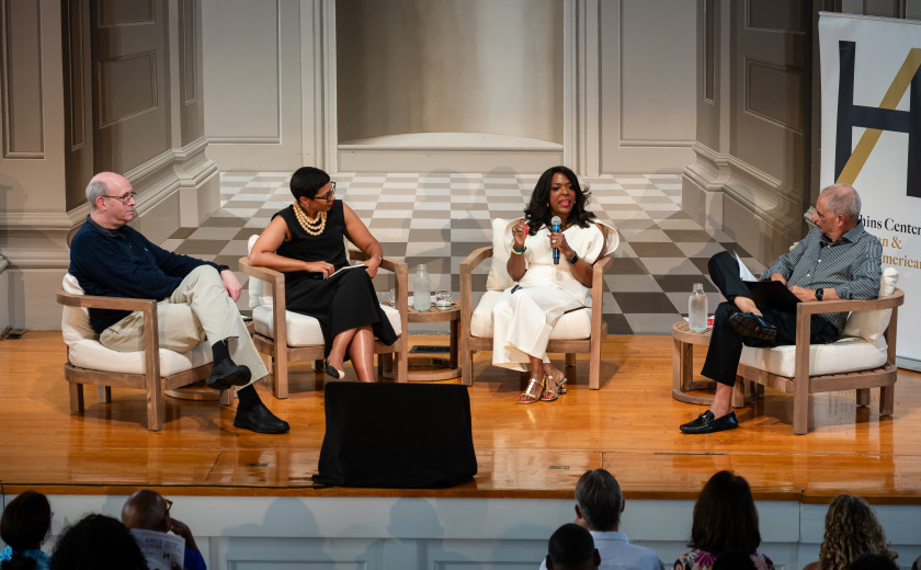 Four panelists seated on armchairs across a wooden stage during a discussion at the Hutchins Forum, with a woman in white holding a microphone in the center while speaking to the group.