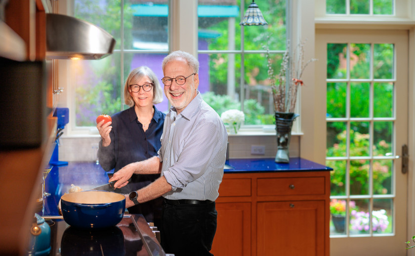 Professor Dan Lieberman and Tonia Prescott in their kitchen. Lieberman stirs a pot on the stove and Prescott holds a tomato.