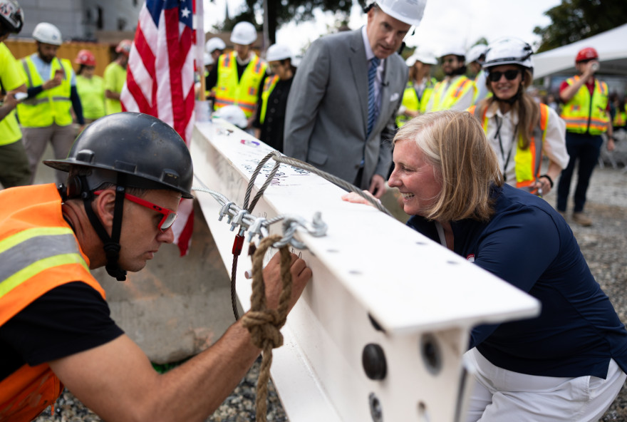 A man in a hard hat and orange vest signs a beam while Dean Hopi Hoekstra looks on