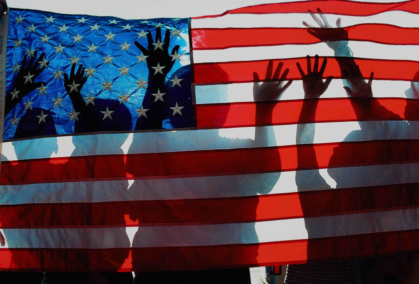 Color image of an American flag with silhouettes of figures with raised arms behind it